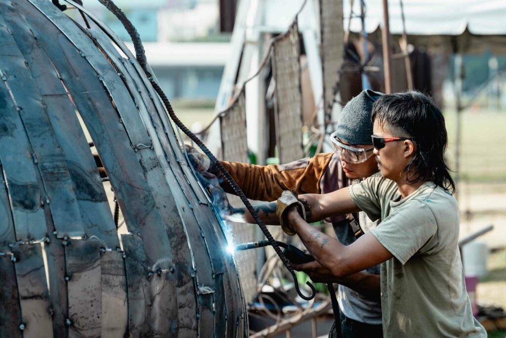 2拼體力也拼創意，藝術家陳俊憲左及助理在焊接火光中打造「戶外現地創作營」鋼雕作品《朝向十方之所》。